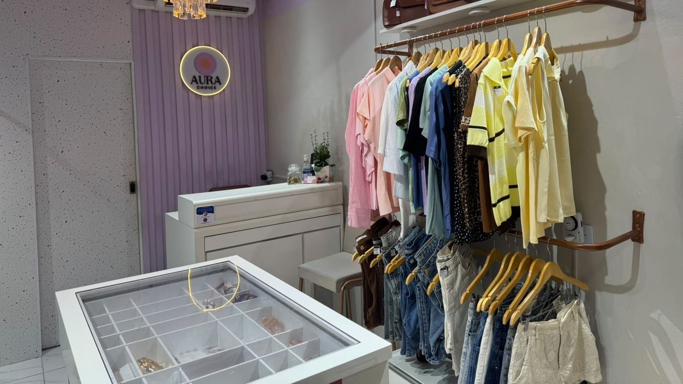 Interior da Aura Choice, com arara de camisas e jeans, balcão de acessórios com o selo de promoção e o logotipo da loja na parede lilás