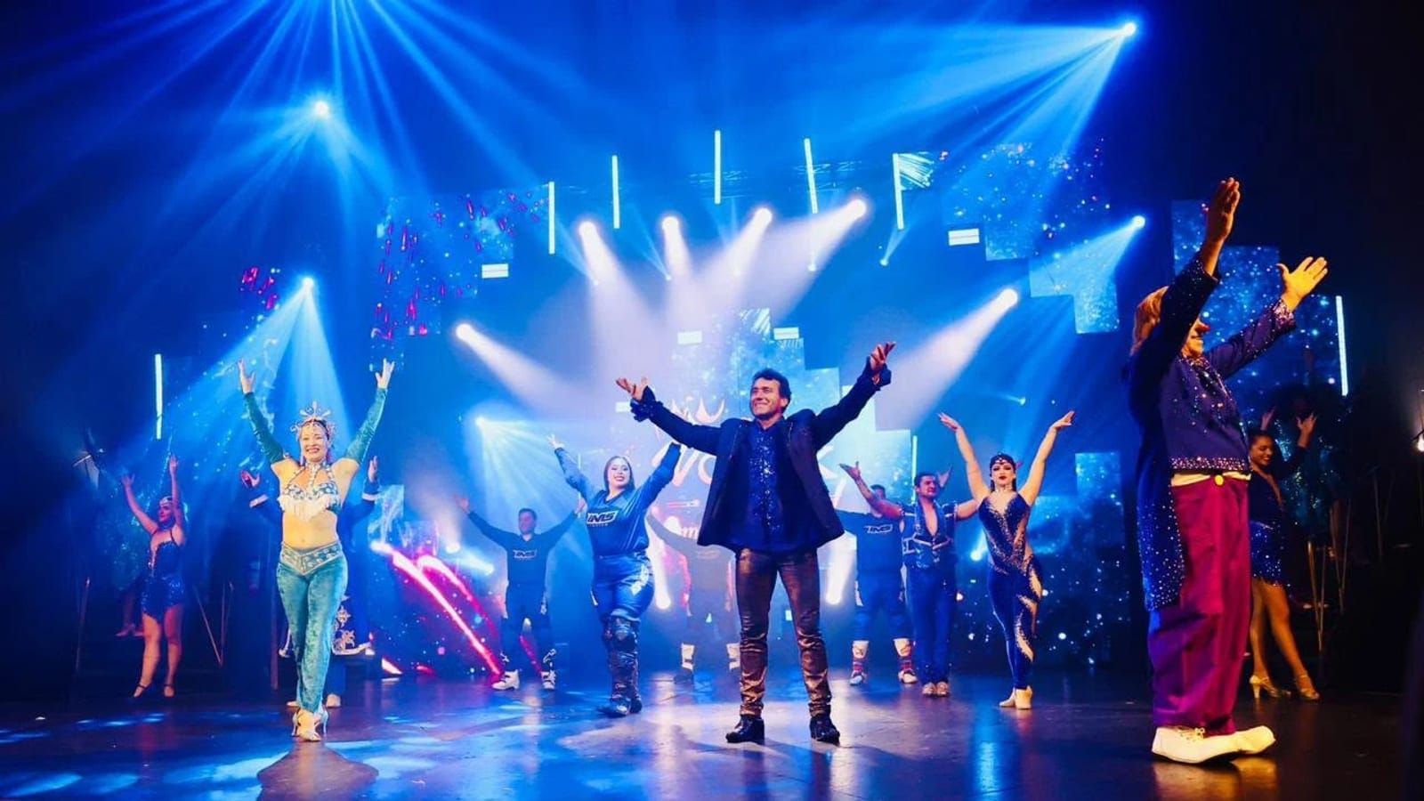 Elenco do Circo Vostok de braços abertos no palco, sob luzes azuis e um painel de LED, no fim do espetáculo Show Emoções