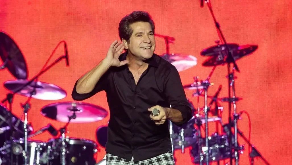 O cantor Daniel sorri com a mão na orelha, de camisa preta, num palco com bateria e telão vermelho