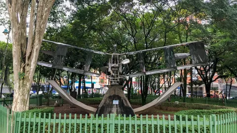 Réplica metálica do 14-Bis cercada por grade verde entre as árvores do Parque Santos Dumont, em São José dos Campos