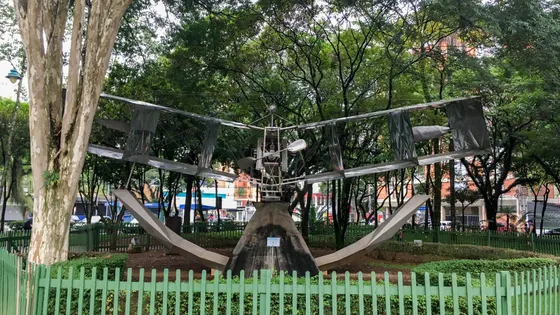 Réplica metálica do 14-Bis cercada por grade verde entre as árvores do Parque Santos Dumont, em São José dos Campos