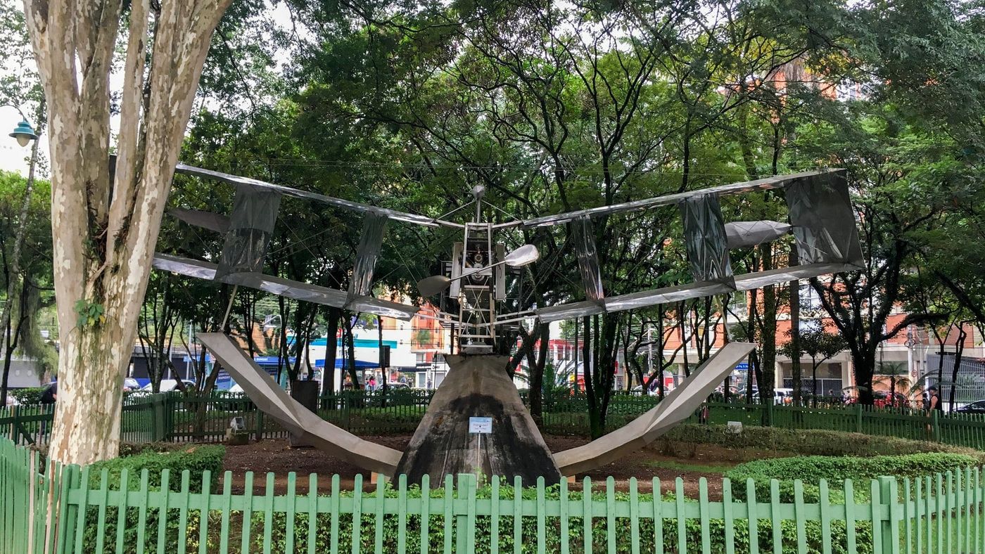 Réplica metálica do 14-Bis cercada por grade verde entre as árvores do Parque Santos Dumont, em São José dos Campos