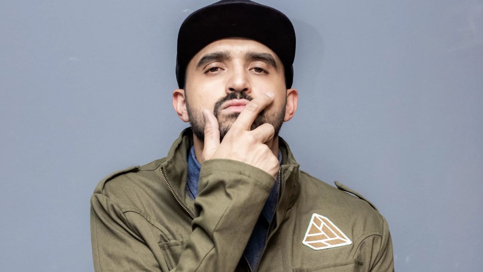 Thiago Ventura, de boné preto e jaqueta verde, com a mão no queixo, em retrato de divulgação sobre fundo cinza