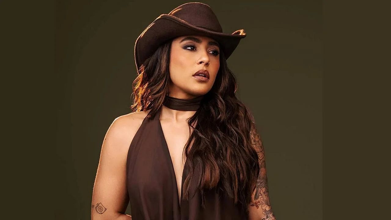 Lauana Prado de chapéu marrom, posando diante de um painel preto com o nome da cantora
