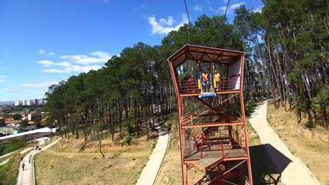 Torre vermelha da tirolesa do Parque Alberto Simões, com visitantes no alto, pinheiros em volta e a cidade ao fundo