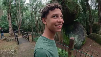 Menino sorrindo na trilha do Parque da Pedra Montada, com as pedras gigantes e a mata ao fundo, em Guararema