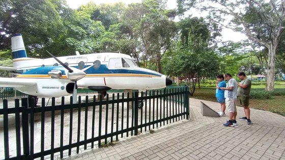 Protótipo do avião Bandeirante, branco com faixas azul e laranja, exposto atrás de uma cerca verde no Parque Santos Dumont, com visitantes ao lado