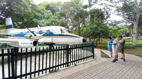 Protótipo do avião Bandeirante, branco com faixas azul e laranja, exposto atrás de uma cerca verde no Parque Santos Dumont, com visitantes ao lado