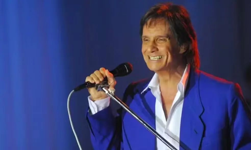 Roberto Carlos sorri ao microfone, de terno azul e camisa branca, diante de um fundo azul